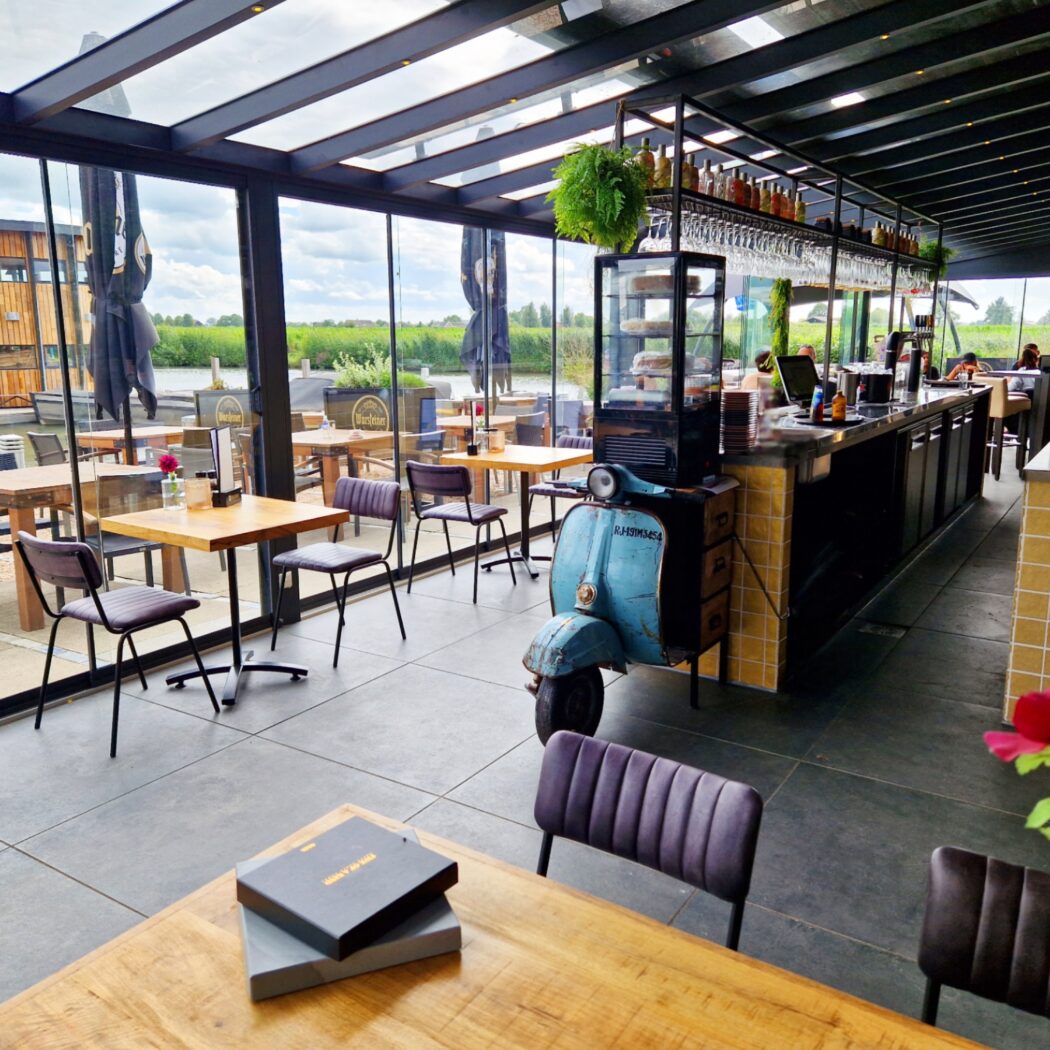 Ruim restaurant met houten tafels, bar, vintage blauwe scooter en glazen wanden met uitzicht op water en houten gebouw.