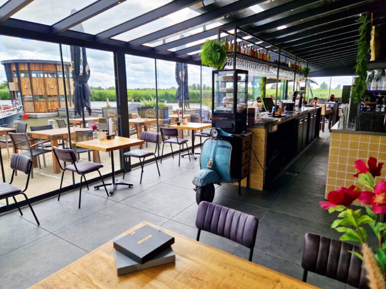 Ruim restaurant met houten tafels, bar, vintage blauwe scooter en glazen wanden met uitzicht op water en houten gebouw.
