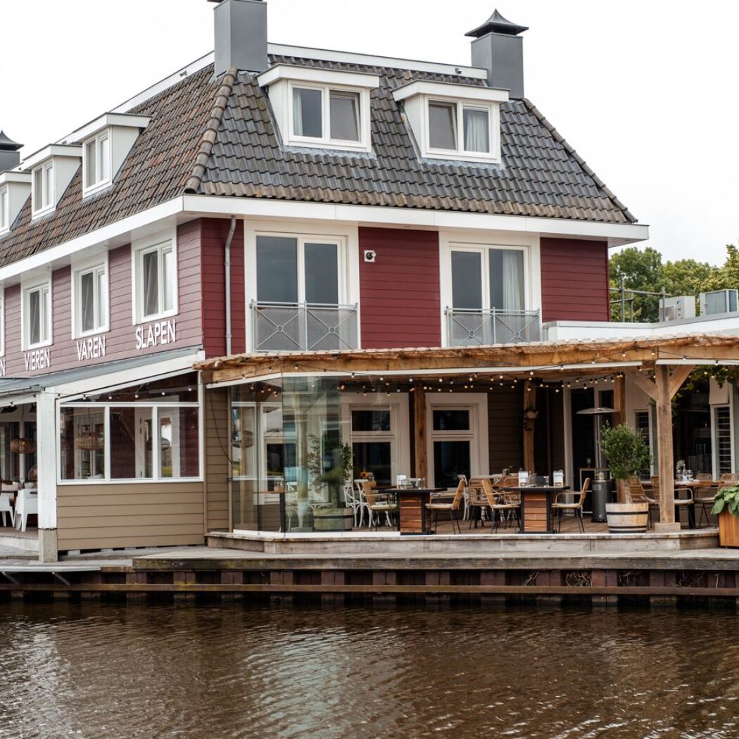 Roodbruin gebouw aan het water met houten terras en tekst DRINKEN VISJES VAREN SLAPEN op de gevel.