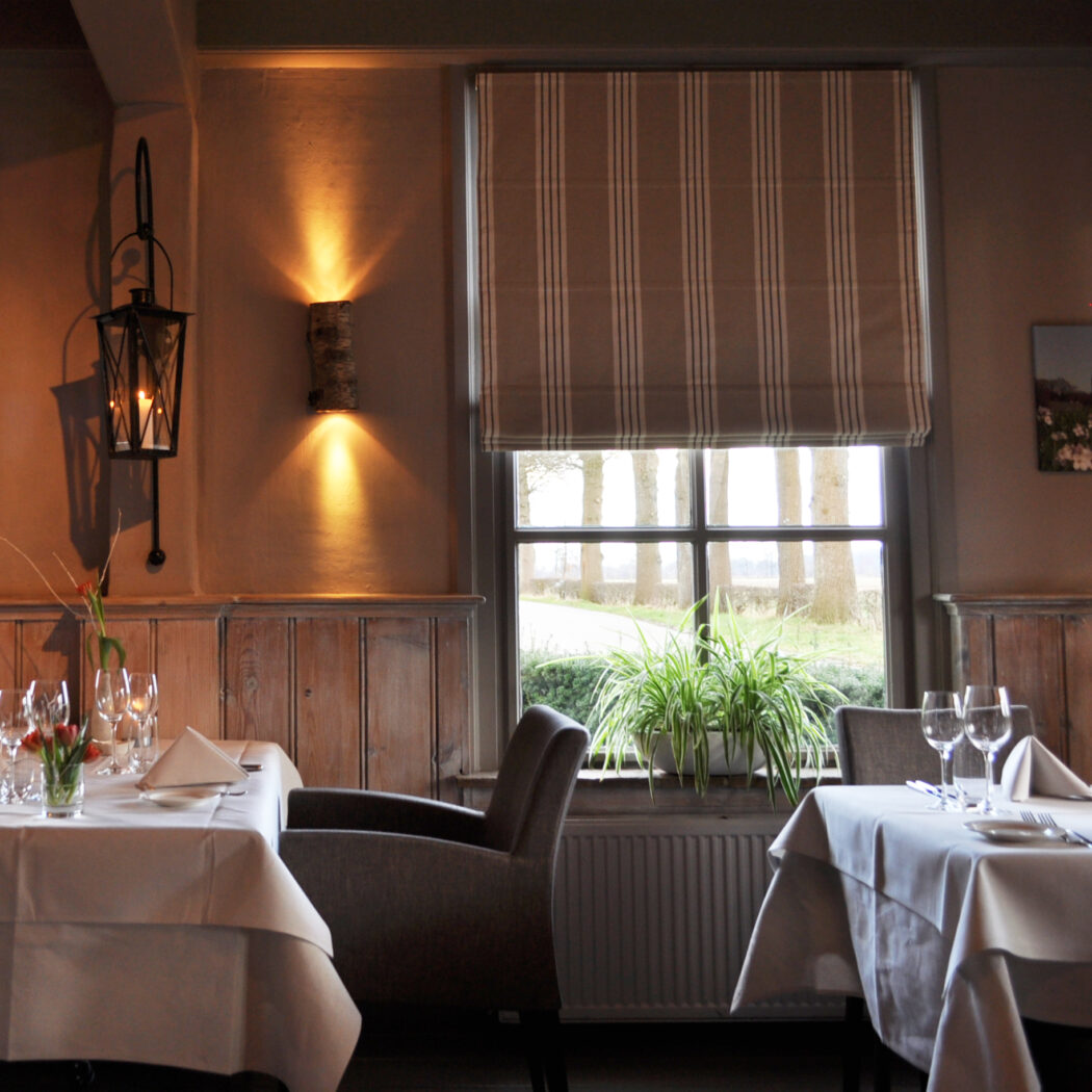 Sfeervol restaurant met gedekte tafels, houten lambrisering, warme verlichting en groen uitzicht.