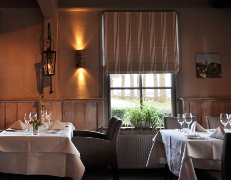 Sfeervol restaurant met gedekte tafels, houten lambrisering, warme verlichting en groen uitzicht.