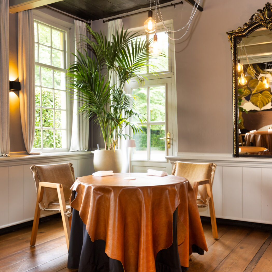 Ronde tafel met bruin leren kleed en twee stoelen in een lichte eetkamer met grote ramen, plant en gouden spiegel.