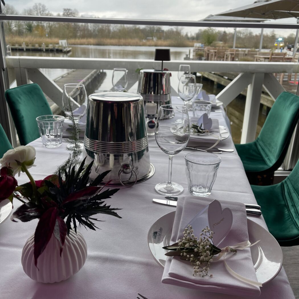 Feestelijk gedekte tafel met witte bloemen, Mevrouw naamkaartje en zilveren koeler, met uitzicht op water en steiger.