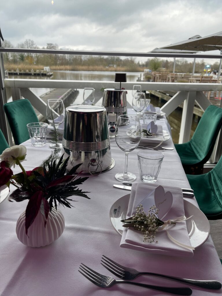 Feestelijk gedekte tafel met witte bloemen, Mevrouw naamkaartje en zilveren koeler, met uitzicht op water en steiger.