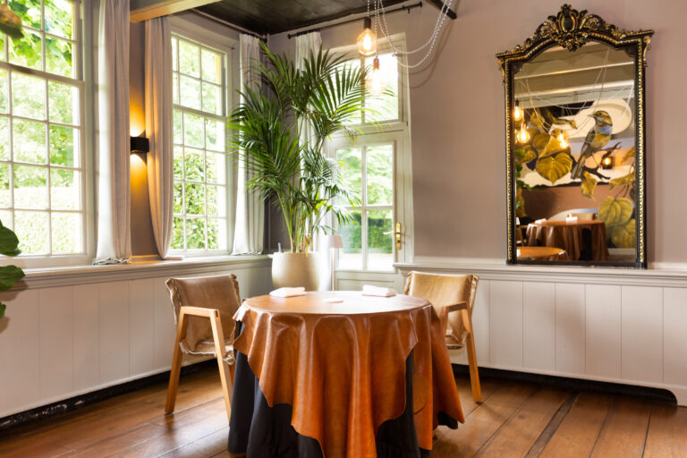Ronde tafel met bruin leren kleed en twee stoelen in een lichte eetkamer met grote ramen, plant en gouden spiegel.