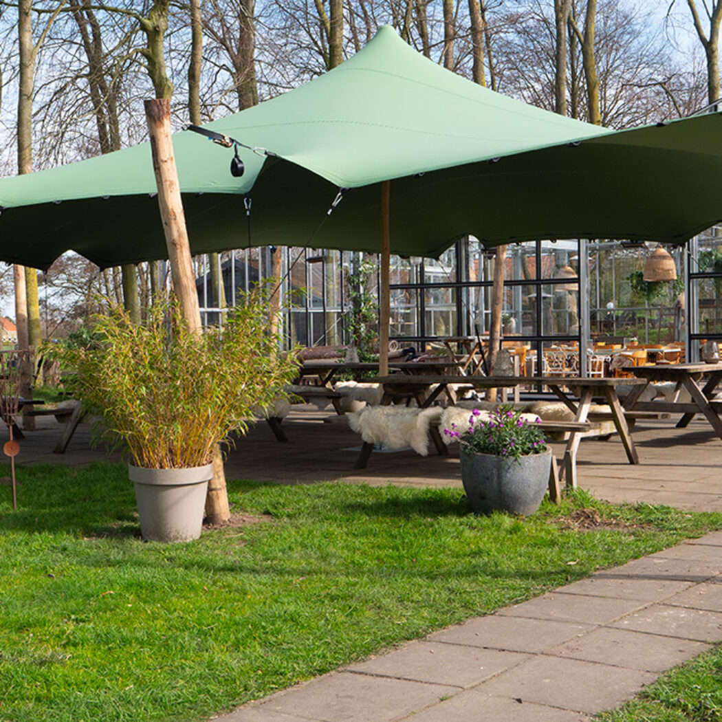 Groene zeiltent boven houten picknicktafels met schapenvachten op een terras bij een glazen kas.
