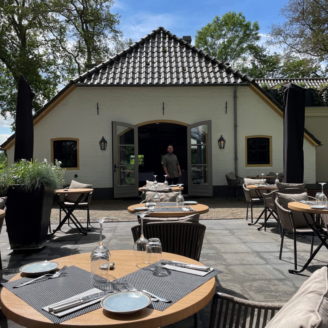 Uitnodigend restaurantterras met gedekte houten tafels, stoelen en een man in de deuropening van een wit gebouw.