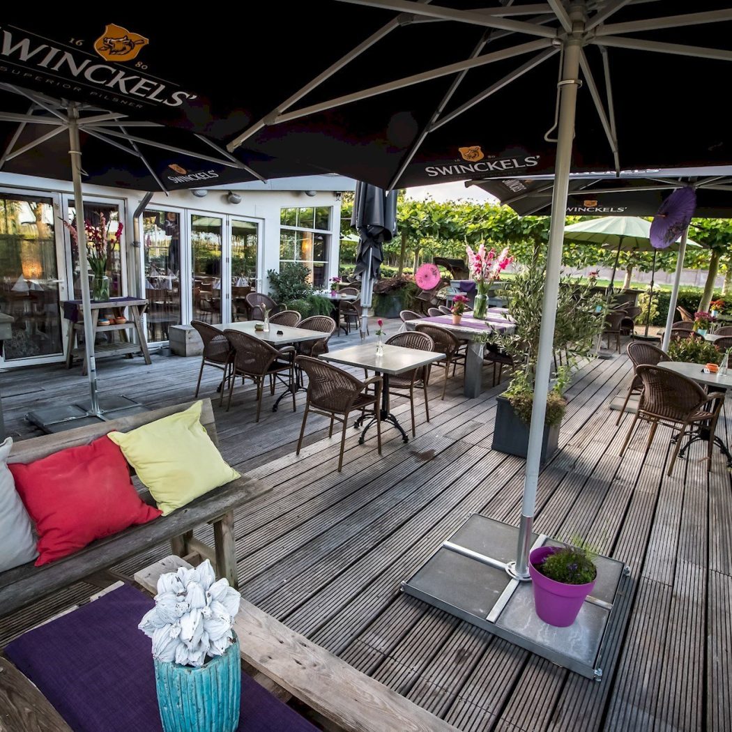 Buitenterras met houten vlonder, tafels, rotan stoelen, zwarte Swinckels parasols en een houten bank met kleurrijke kussens.