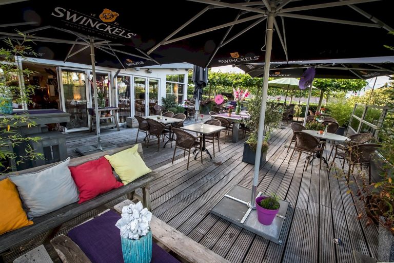 Buitenterras met houten vlonder, tafels, rotan stoelen, zwarte Swinckels parasols en een houten bank met kleurrijke kussens.
