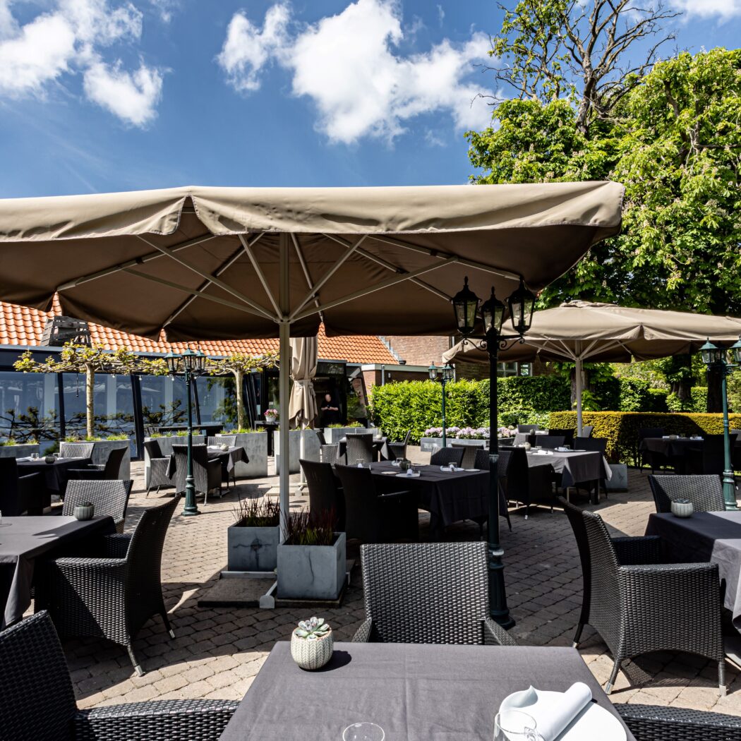 Ruim restaurantterras met veel tafels, donkere stoelen en grote beigekleurige parasols op een zonnige dag.