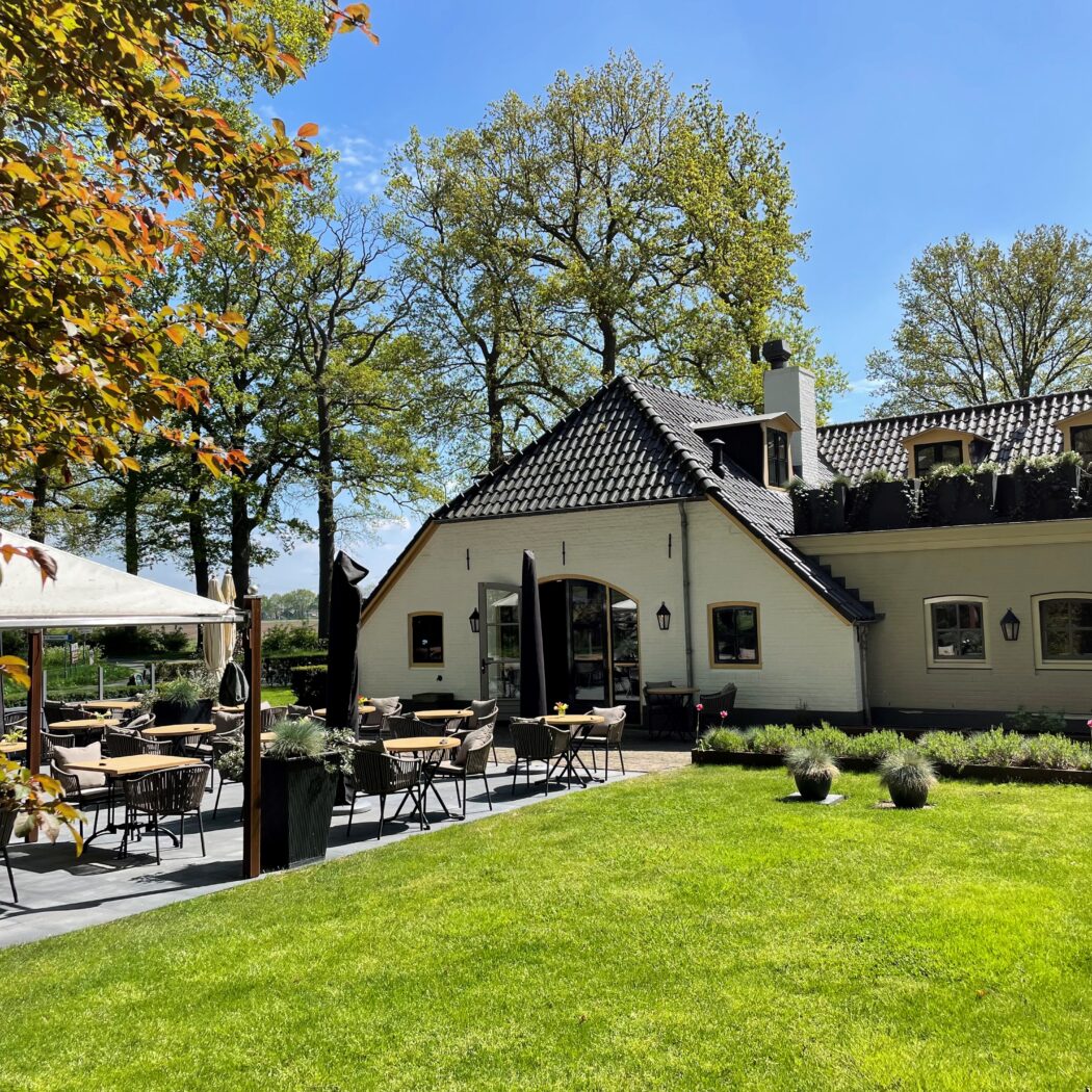 Zonnig beeld van een landelijk gebouw met een buitenterras, omgeven door een groen gazon en hoge bomen.