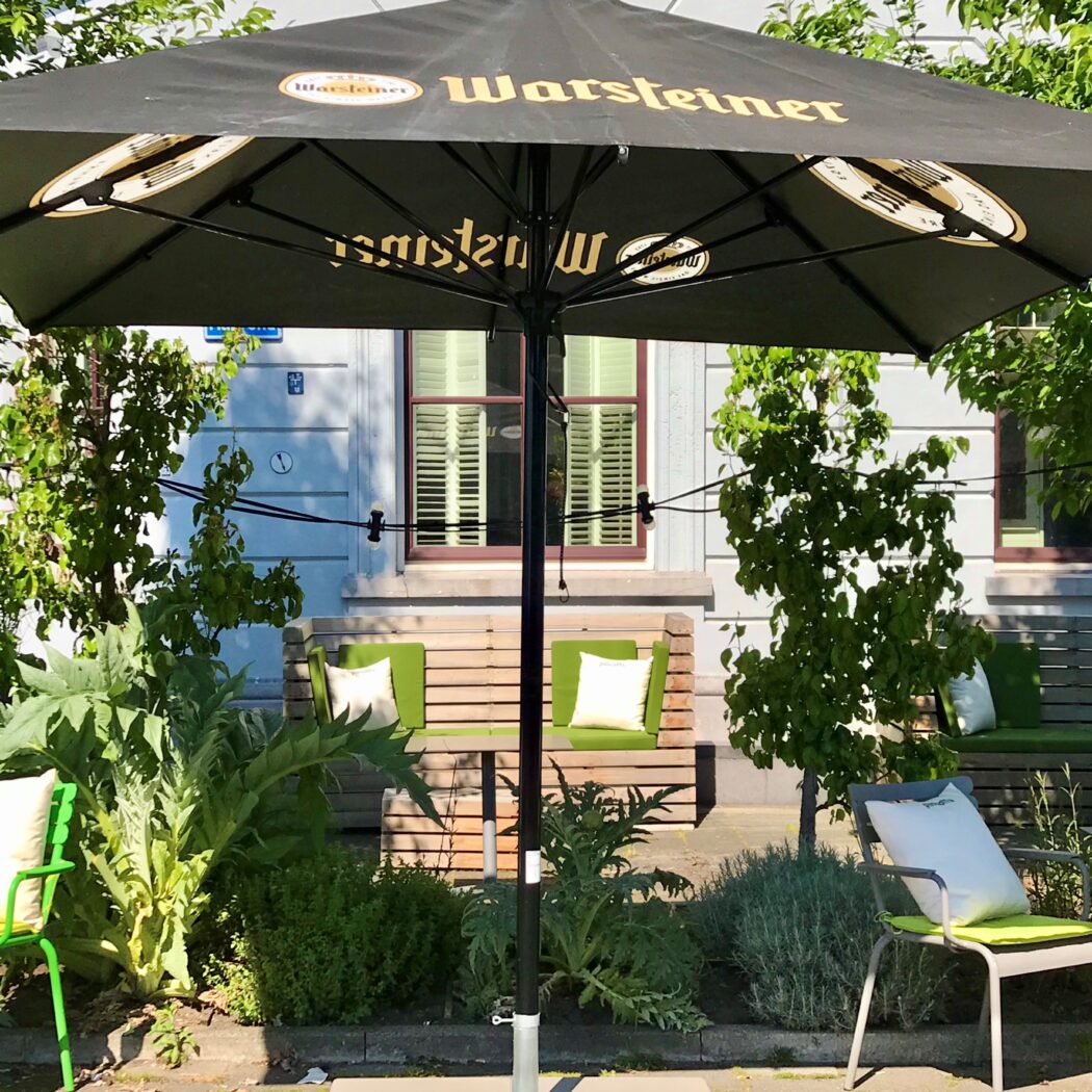 Zwarte Warsteiner parasol boven een zonnig terras met groene stoelen, banken en weelderige planten.