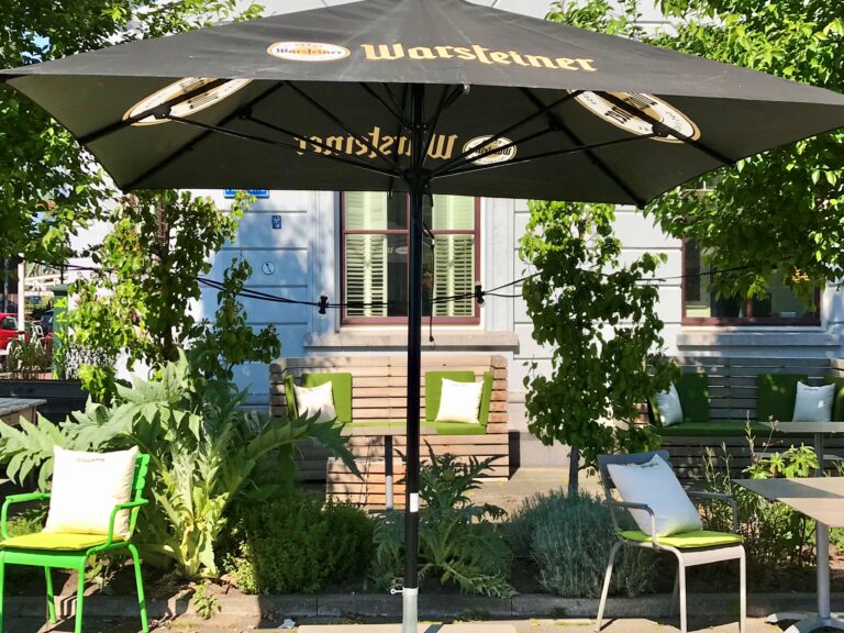Zwarte Warsteiner parasol boven een zonnig terras met groene stoelen, banken en weelderige planten.