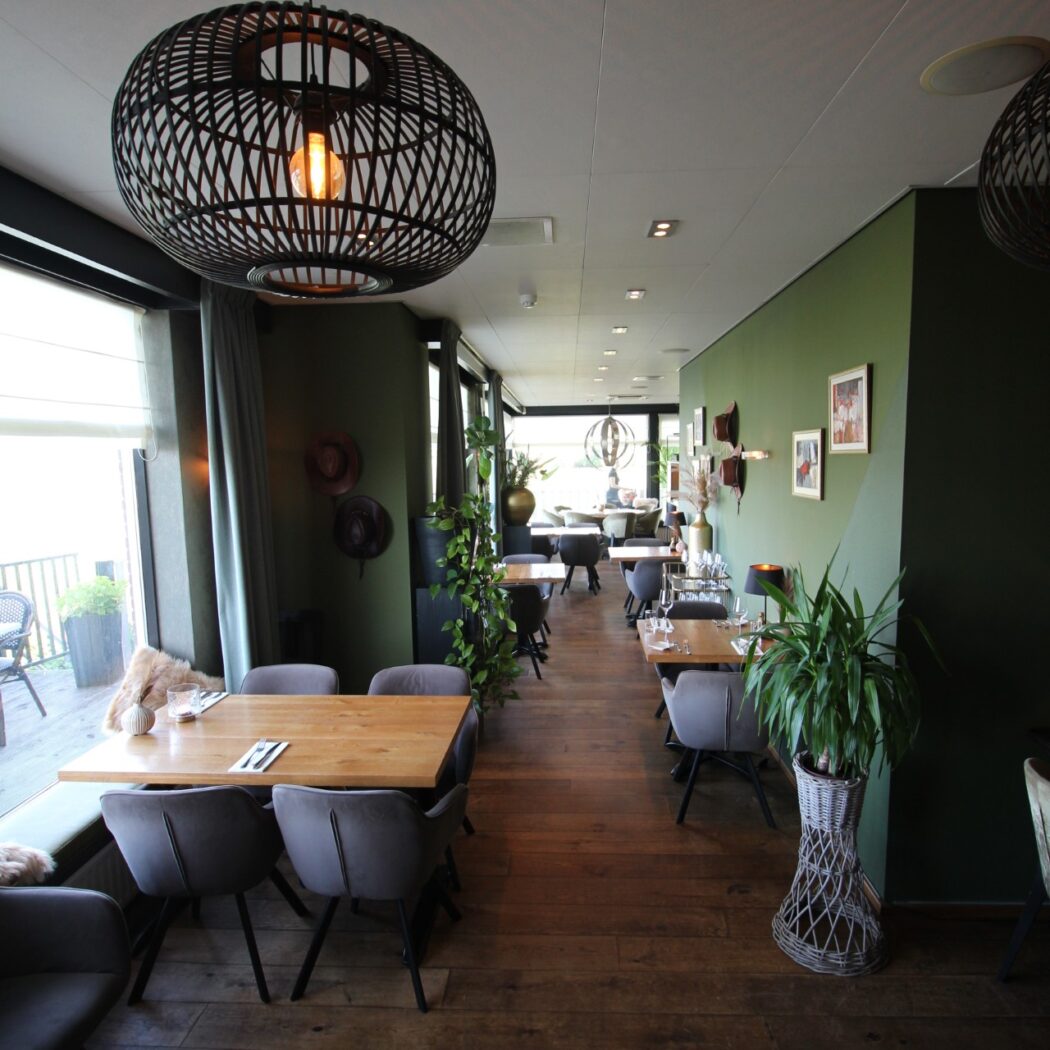 Sfeervol restaurant met donkergroene muren, houten vloer, zwarte geweven hanglamp en gedekte tafels bij het raam.