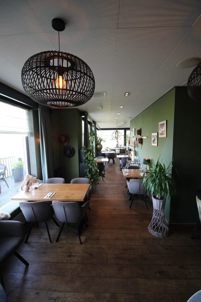 Sfeervol restaurant met donkergroene muren, houten vloer, zwarte geweven hanglamp en gedekte tafels bij het raam.