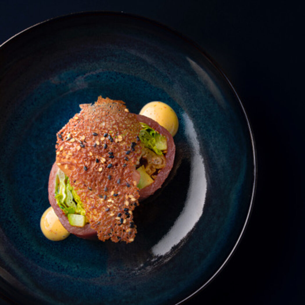 Kunstig opgemaakt gerecht met paarsrode basis, groene groenten, krokante chip met sesam en gele saus in een diepblauwe kom.
