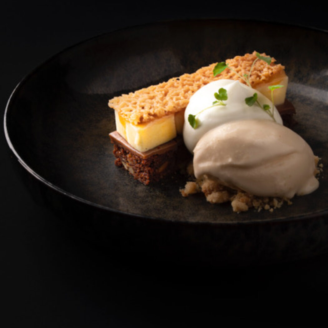 Gelaagd dessert met cheesecake, krokante crumble, witte room en lichtbruin ijs op een donker bord.