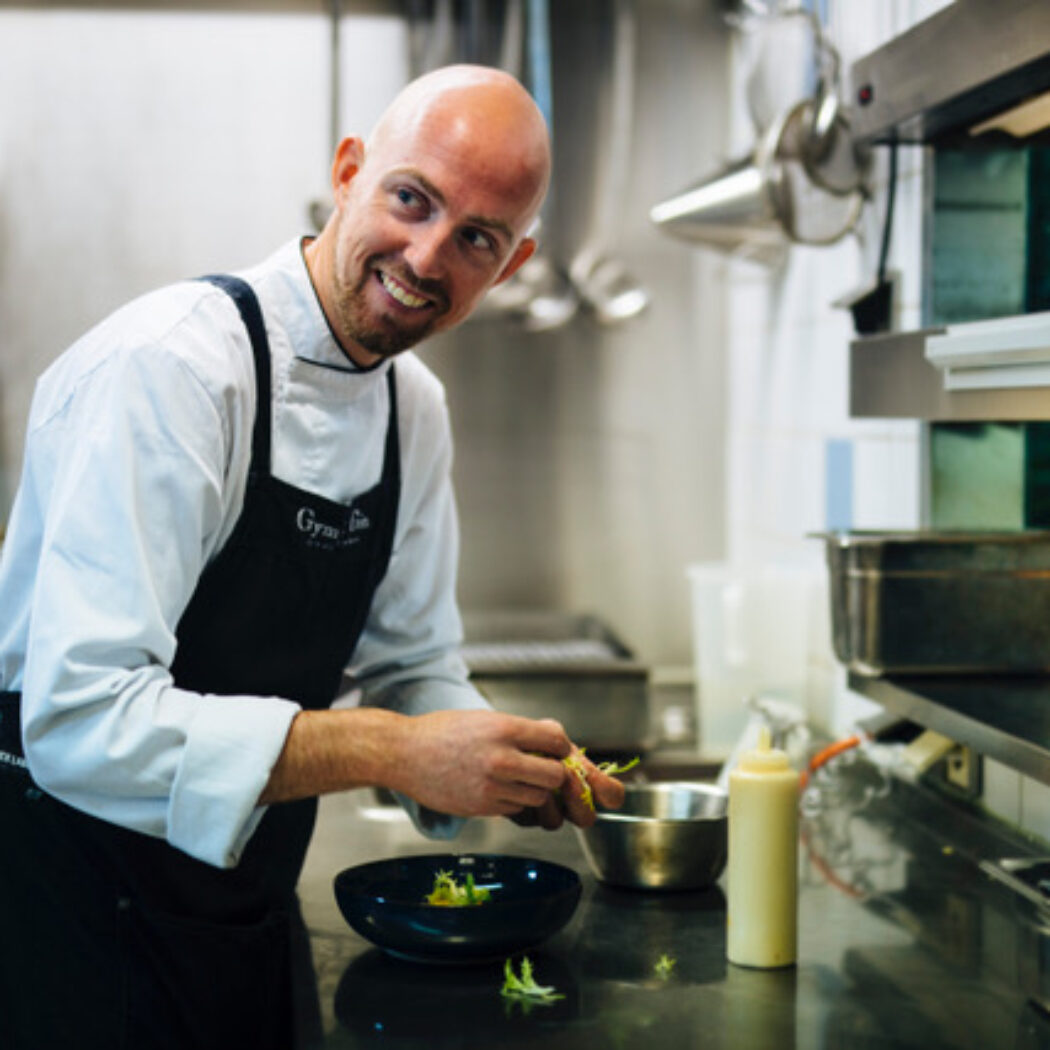 Lachende, kale chef-kok met Gym Life schort garneert een gerecht in een professionele keuken.