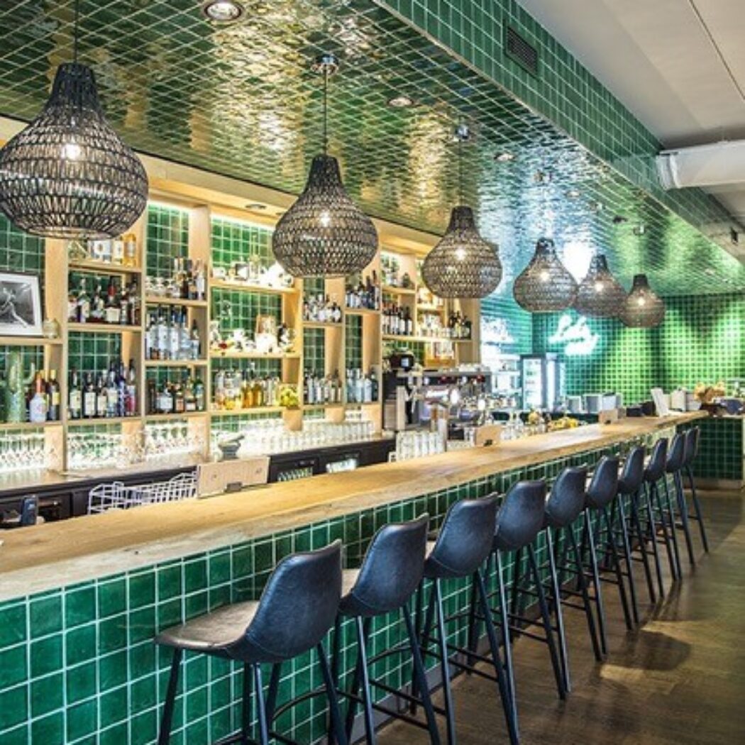 Modern bar interieur met houten toonbank, groene tegels op bar en plafond, zwarte barkrukken en geweven hanglampen.