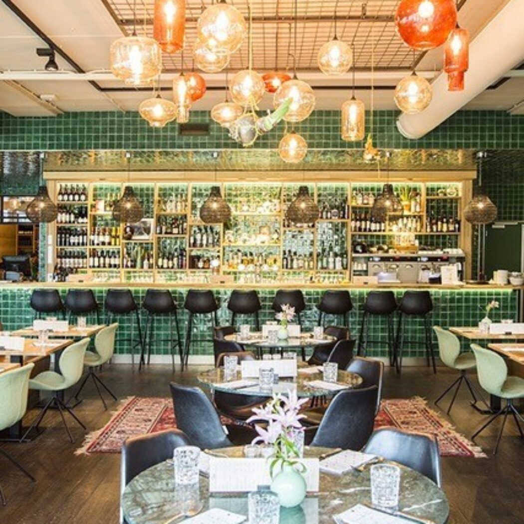 Sfeervol restaurantinterieur met groene tegelwand achter de bar, marmeren tafels en diverse hanglampen.
