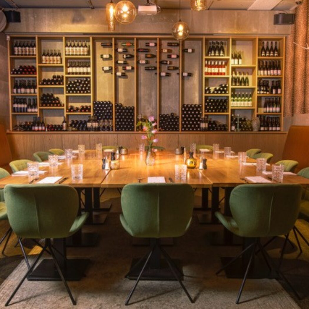Gedekte lange houten eettafel met groene stoelen in een sfeervol restaurant met wijnmuur.