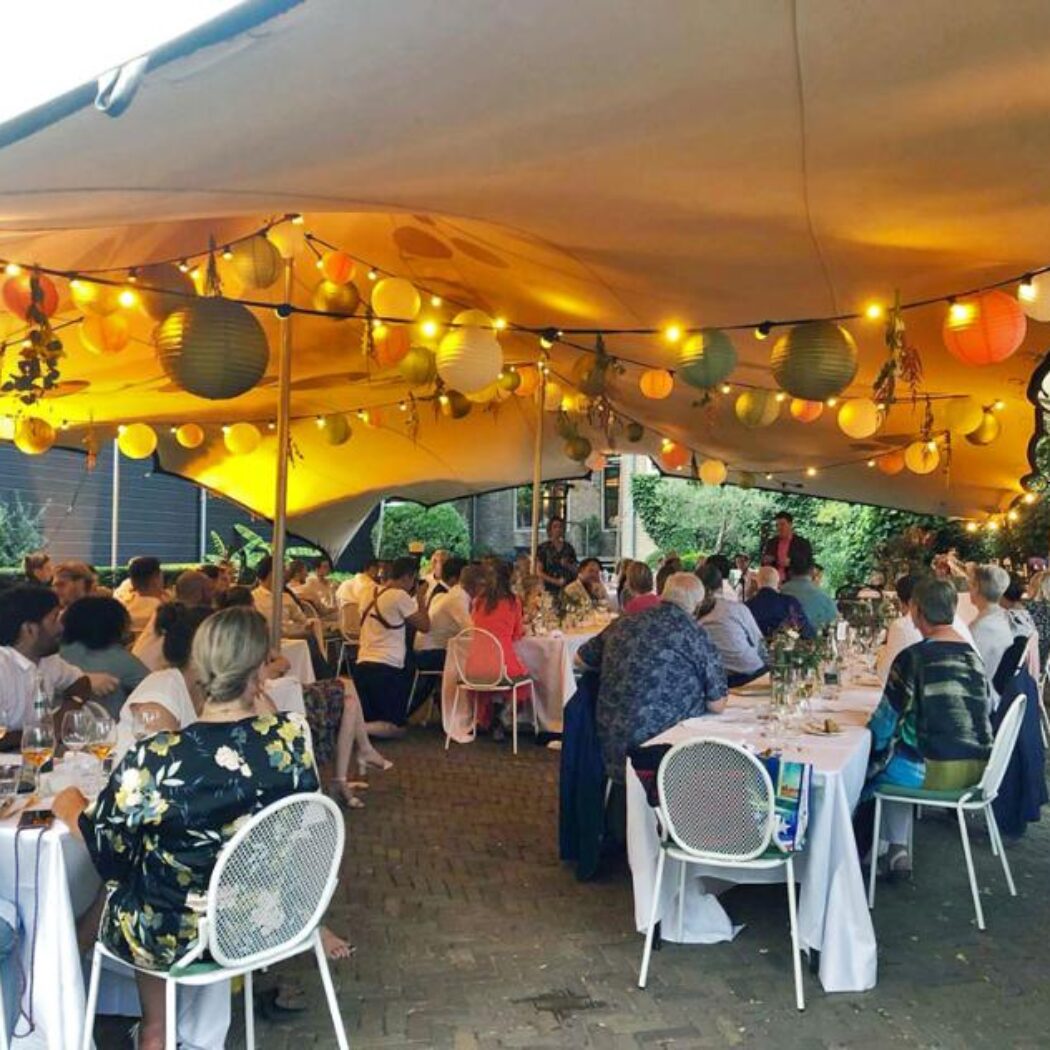 Gasten dineren buiten onder een tent versierd met kleurrijke lampionnen en sfeervolle lichtslingers.