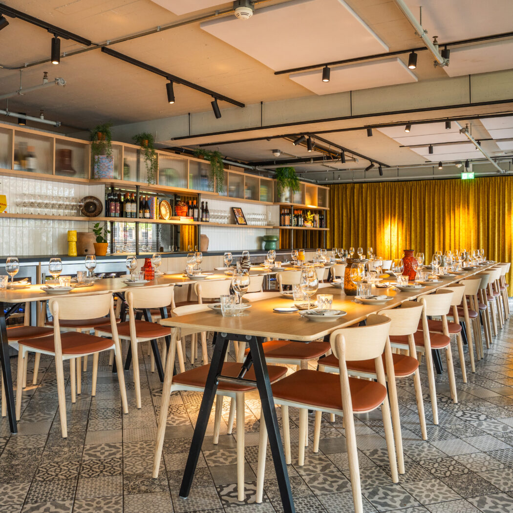 Lange, modern gedekte tafels en houten stoelen in een sfeervolle eetruimte met bar en gele gordijnen.