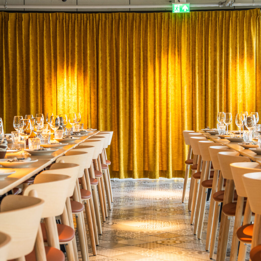 Lange gedekte tafels met stoelen in een moderne eetzaal met een goudgeel fluwelen gordijn.