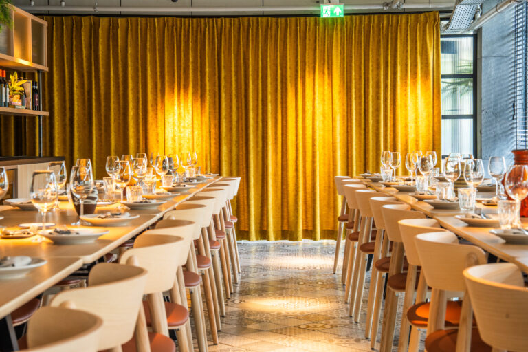Lange gedekte tafels met stoelen in een moderne eetzaal met een goudgeel fluwelen gordijn.