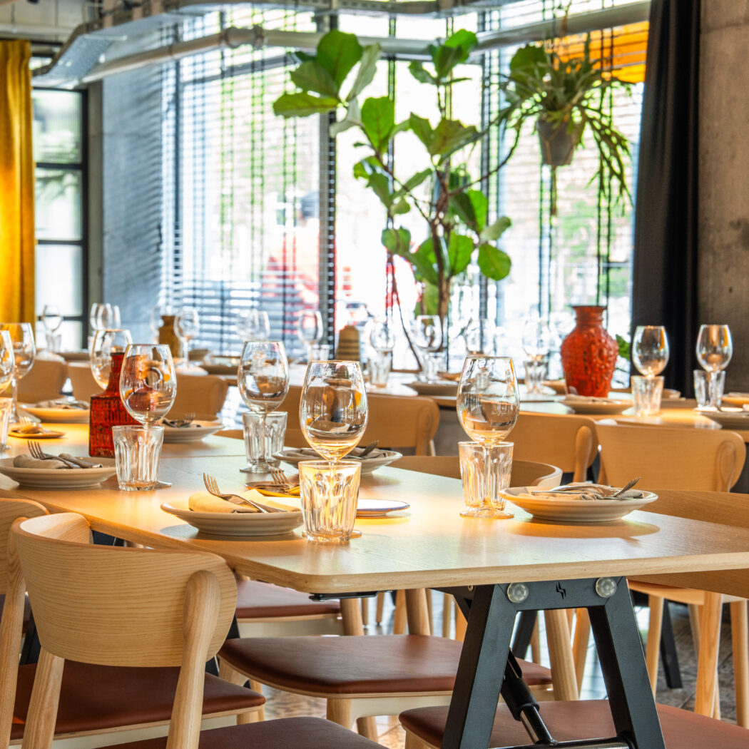 Gedekte houten tafels en stoelen in een modern restaurant, klaar voor gasten, met planten en een rode vaas.