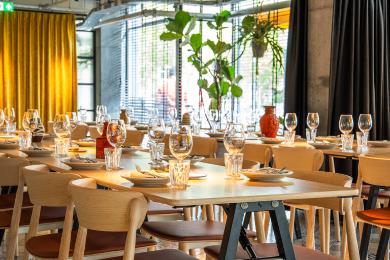 Gedekte houten tafels en stoelen in een modern restaurant, klaar voor gasten, met planten en een rode vaas.