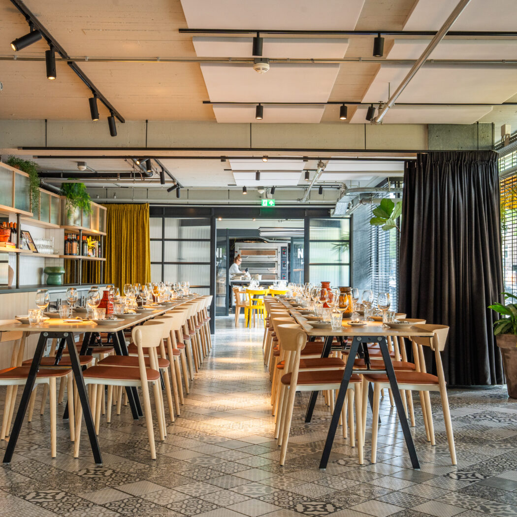 Moderne restaurantzaal met twee lange gedekte houten tafels, patroontegels en een bar met flessen en planten.