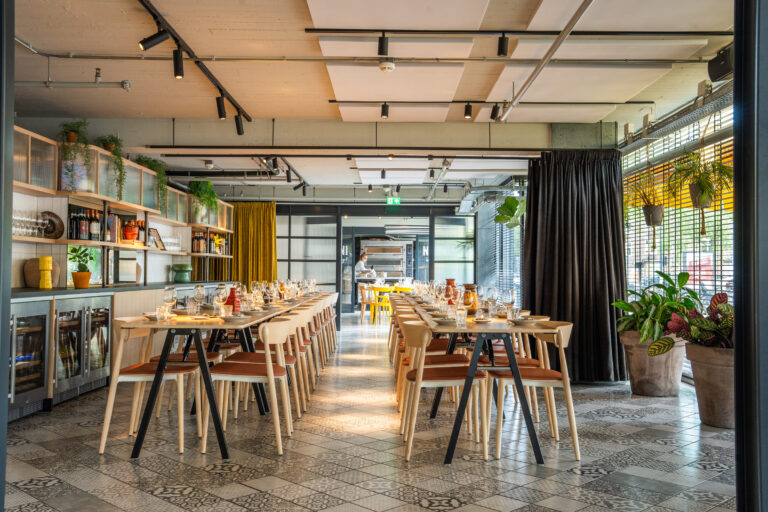Moderne restaurantzaal met twee lange gedekte houten tafels, patroontegels en een bar met flessen en planten.