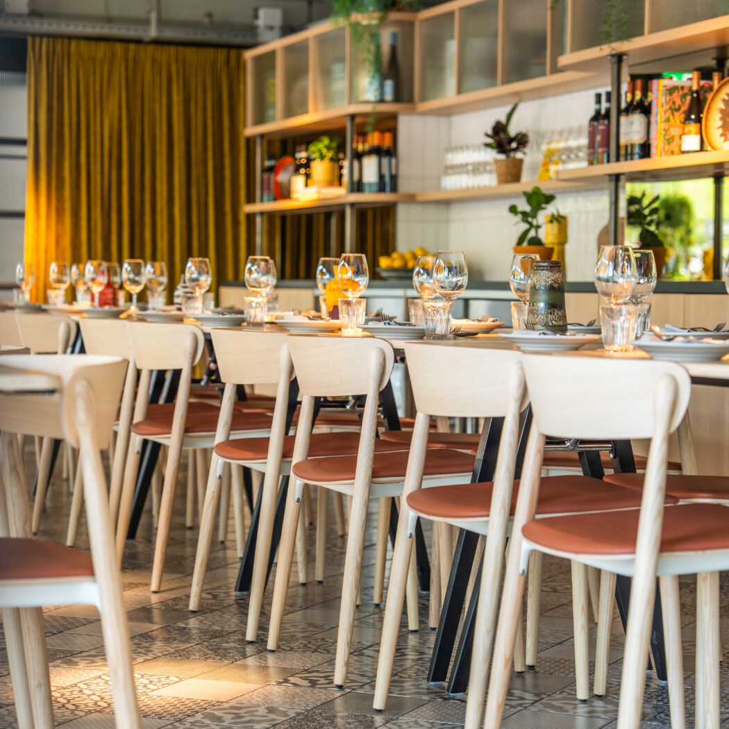 Lange restauranttafel met gedekt servies en licht houten stoelen met oranje zittingen, voor een okergeel gordijn.