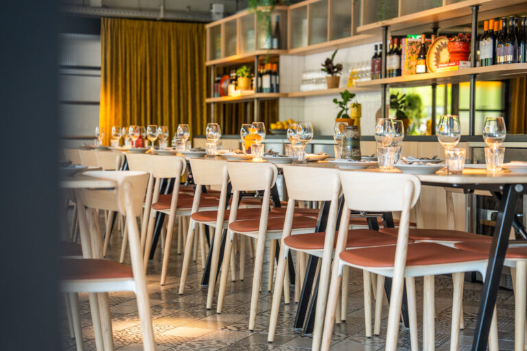 Lange restauranttafel met gedekt servies en licht houten stoelen met oranje zittingen, voor een okergeel gordijn.