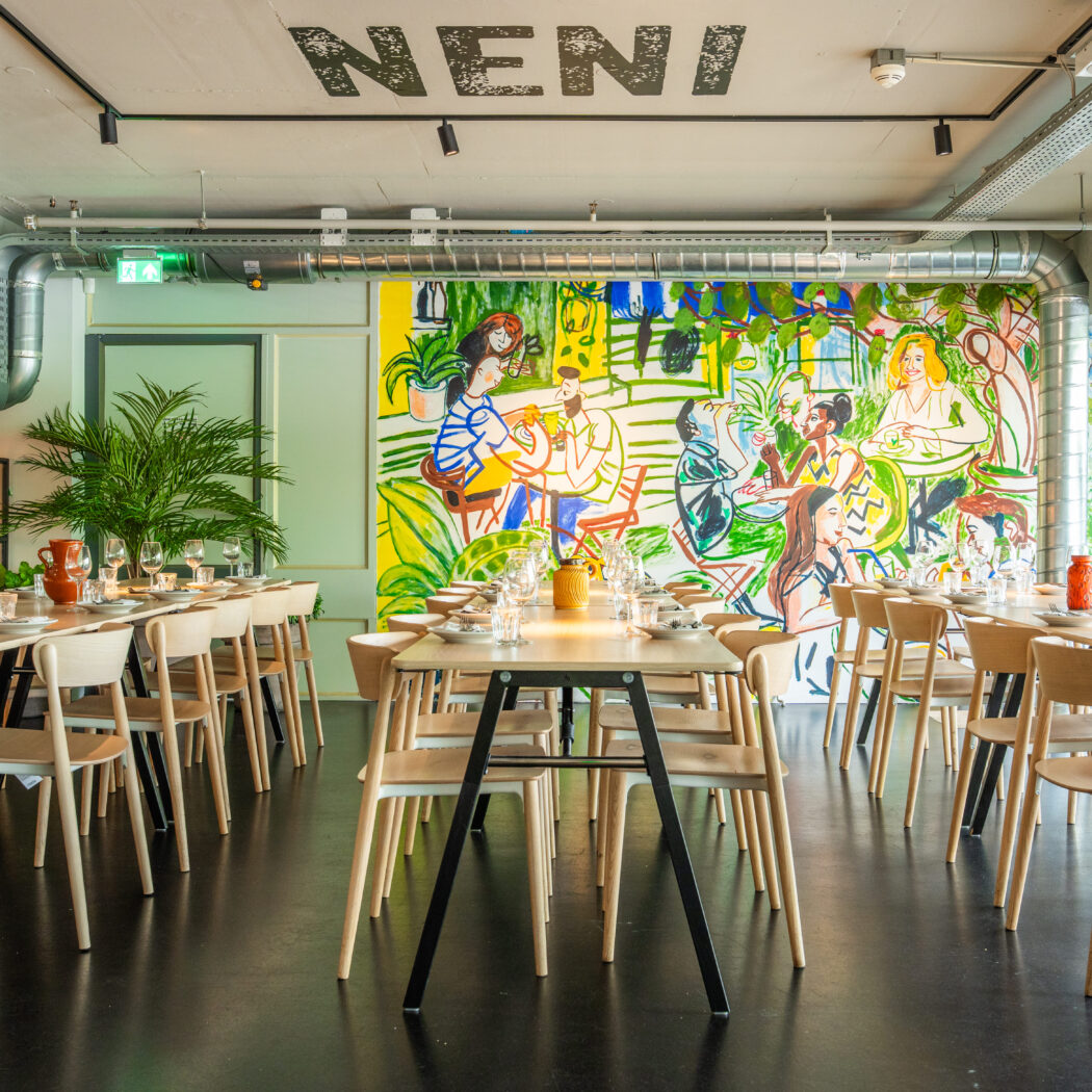 Interieur van restaurant NENI met gedekte tafels, houten stoelen en een grote kleurrijke muurschildering van mensen.