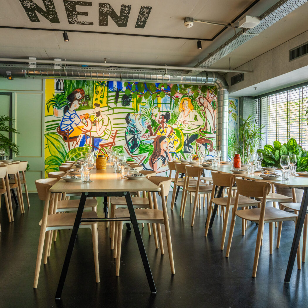 Moderne eetzaal in restaurant NENI met lange, gedekte tafels, houten stoelen en een kleurrijke muurschildering.