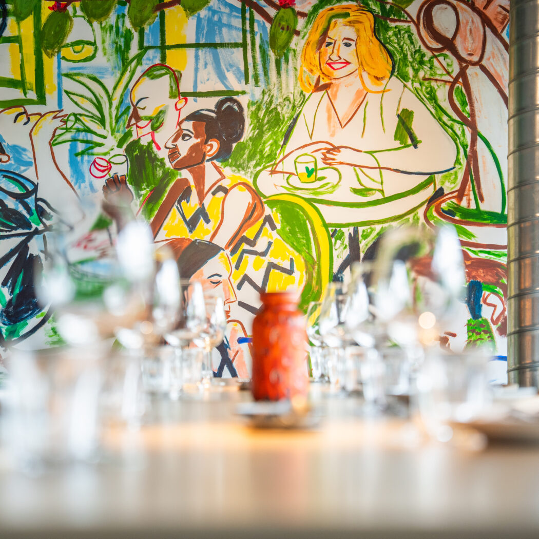 Kleurrijke muurschildering van figuren en planten in een restaurant, met een tafel vol glazen en een oranje vaas.
