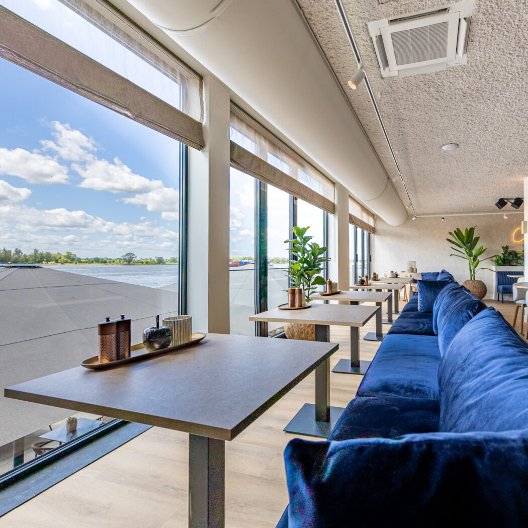 Lichte restaurantruimte met blauwe fluwelen bank, tafels en weids uitzicht op het water.
