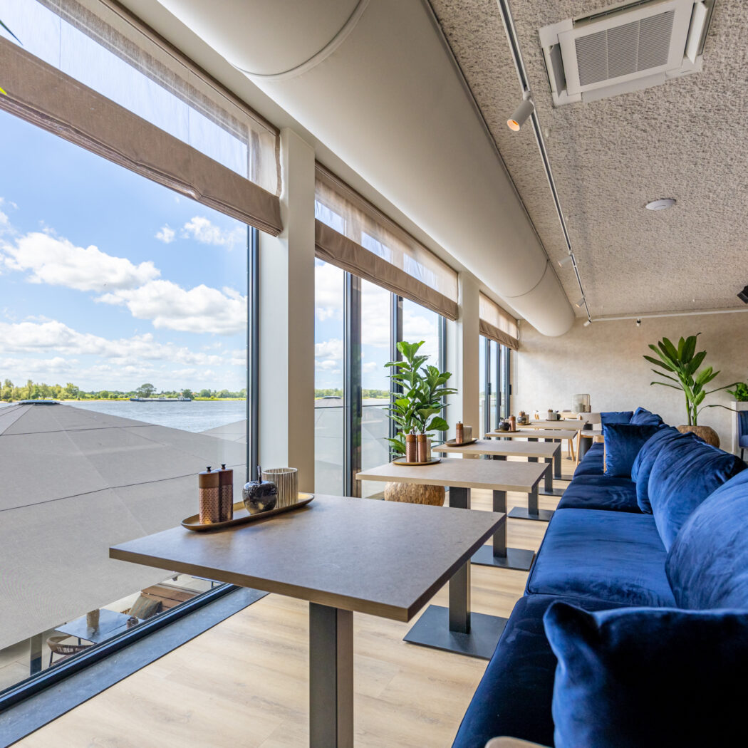 Modern restaurantinterieur met lange blauwe fluwelen bank, tafels en panoramisch uitzicht op water.