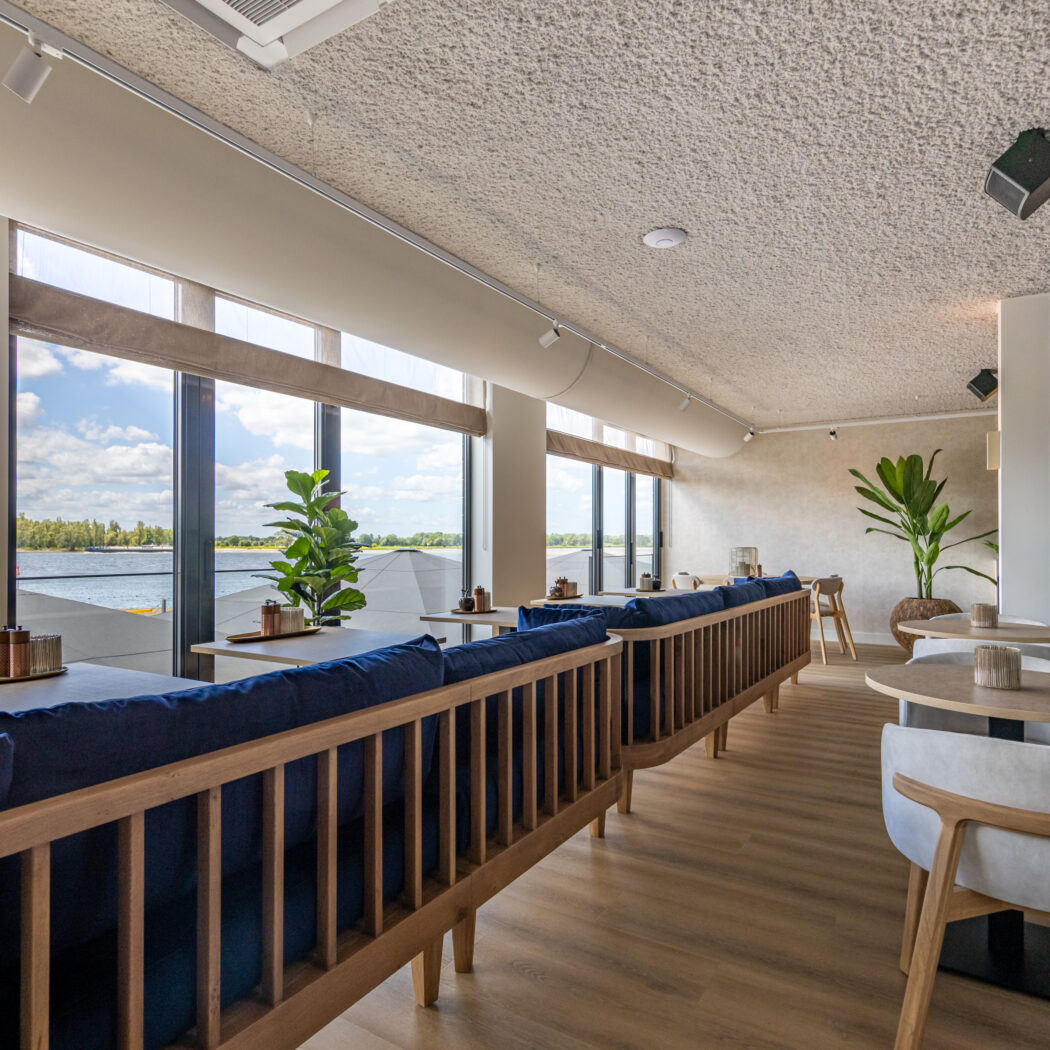 Licht, modern restaurant met blauwe banken, houten meubels en panoramisch uitzicht op water.