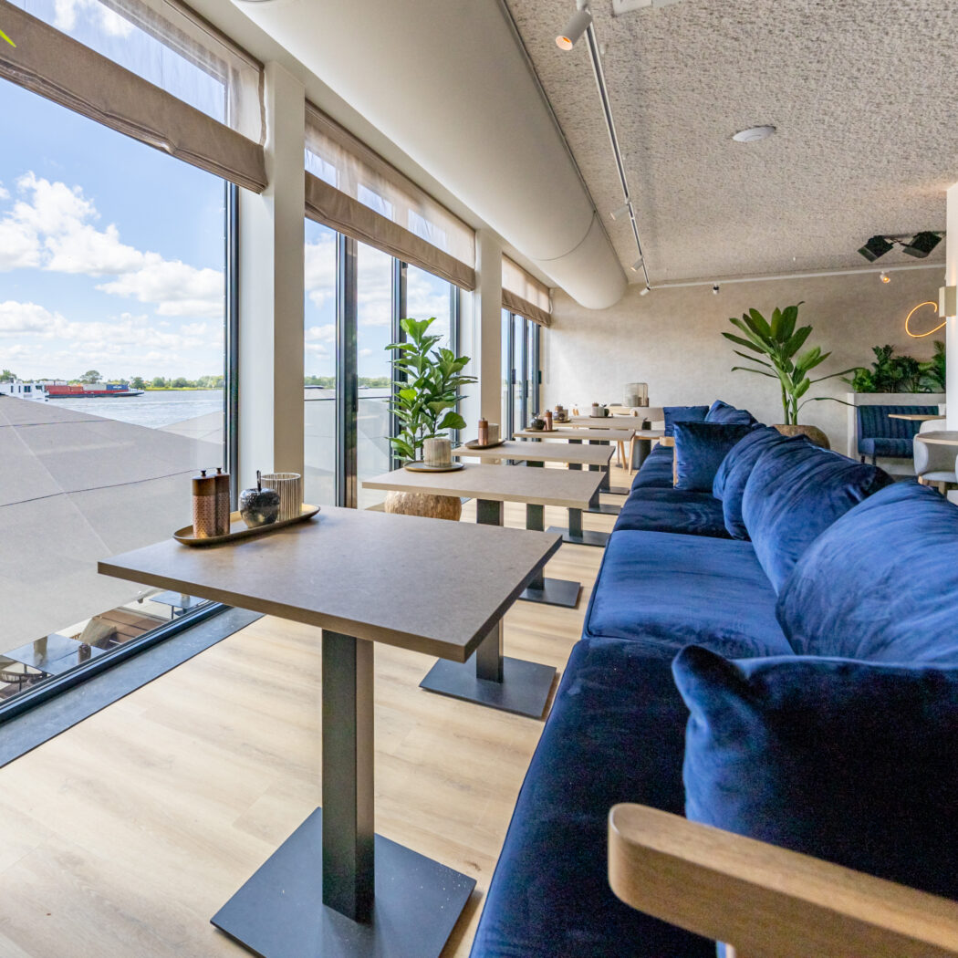 Licht restaurant met lange blauwe velours bank, kleine tafels en uitzicht op water en boten.