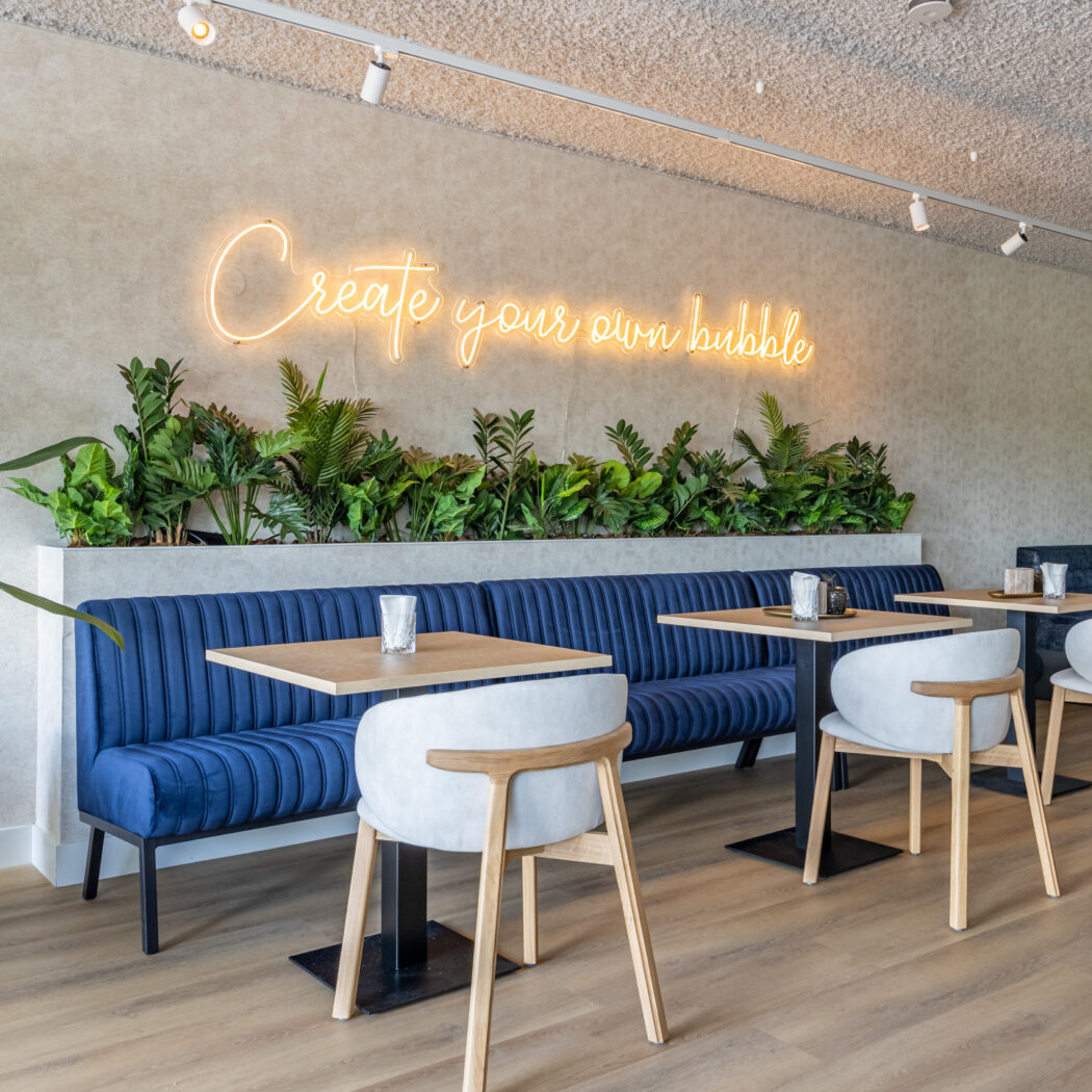 Modern café interieur met lange blauwe zitbank, houten tafels, witte stoelen en neonbord ‘Create your own bubble’ boven groene planten.
