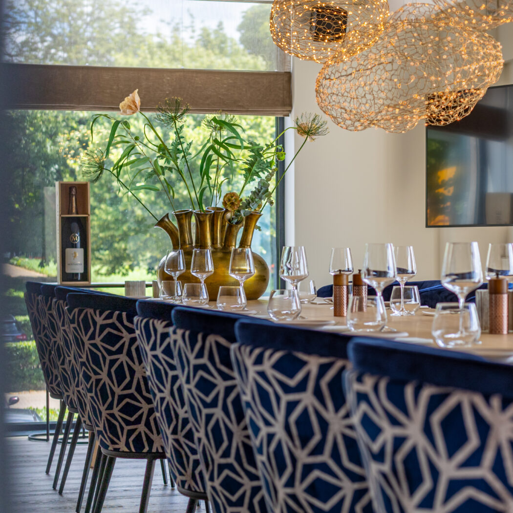 Lange houten vergadertafel met blauw-wit geometrische stoelen, bronzen vaas, fles in kist, en gouden designlampen.