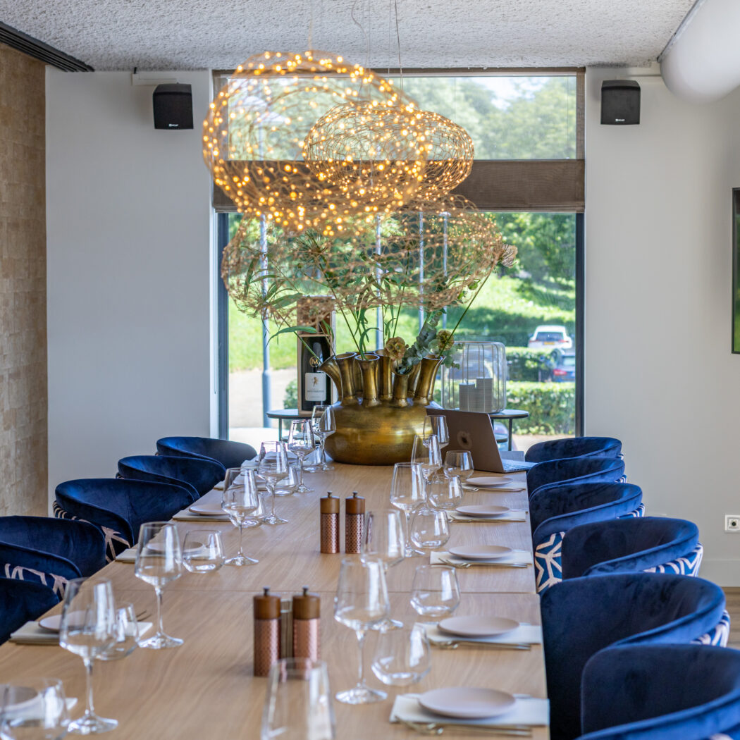 Moderne dinerruimte met lange gedekte houten tafel, blauwe fluwelen stoelen, gouden vaas en bolvormige hanglampen.