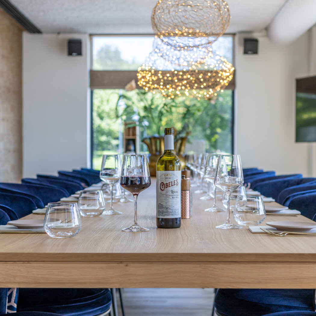 Lange houten tafel met gedekte plaatsen, omringd door blauwe fluwelen stoelen, en een fles rode Cibeles Rioja wijn.