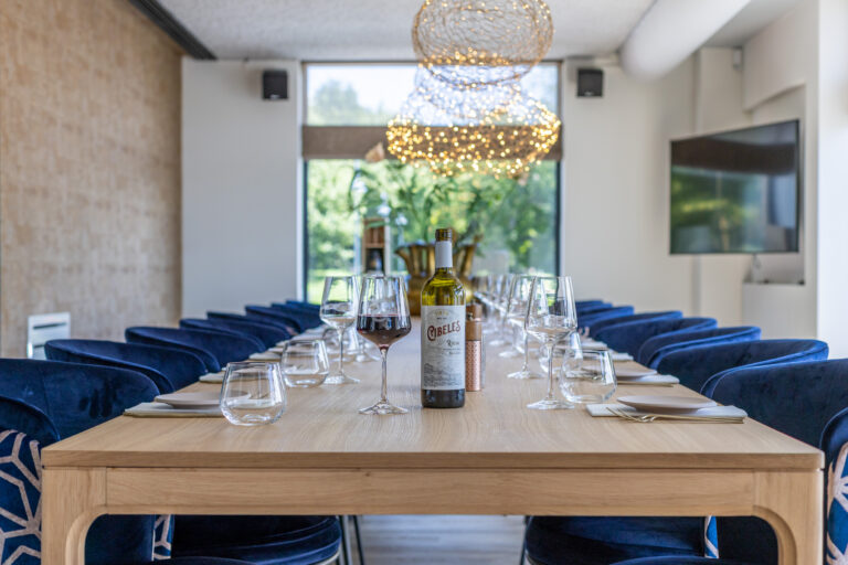 Lange houten tafel met gedekte plaatsen, omringd door blauwe fluwelen stoelen, en een fles rode Cibeles Rioja wijn.