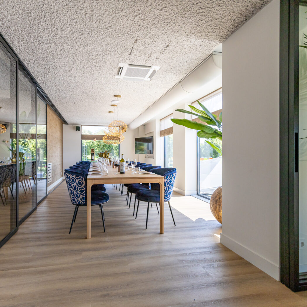Modern ingerichte privéruimte met lange houten eettafel, blauwe stoelen, glazen wanden en veel groen.