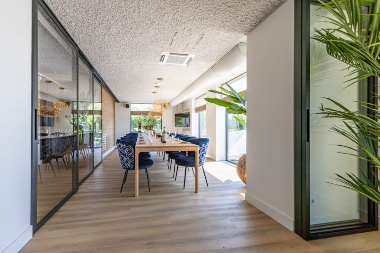 Modern ingerichte privéruimte met lange houten eettafel, blauwe stoelen, glazen wanden en veel groen.