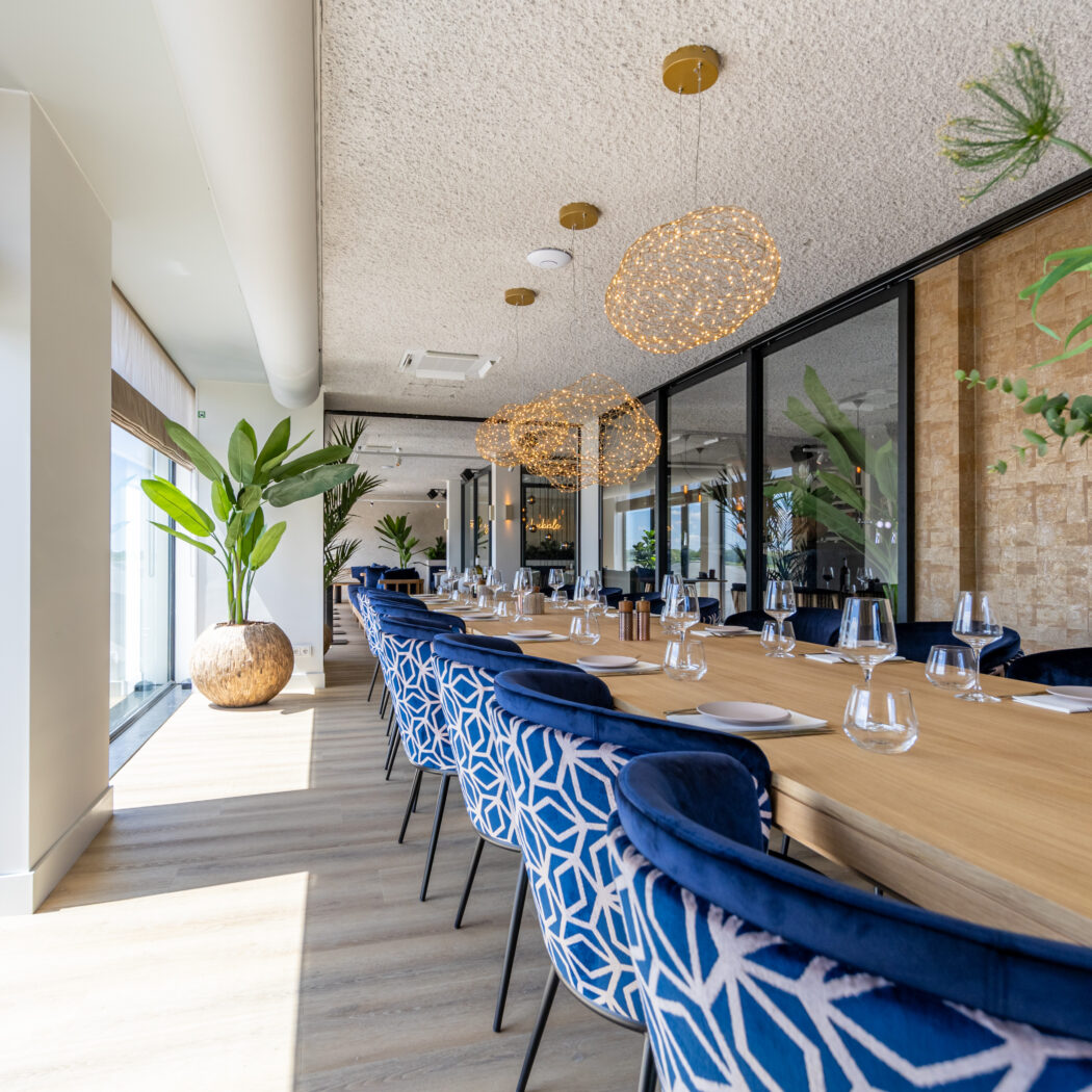 Lange houten eettafel met gedekte plaatsen en blauwe fluwelen stoelen in een moderne zaal met ramen en gouden hanglampen.